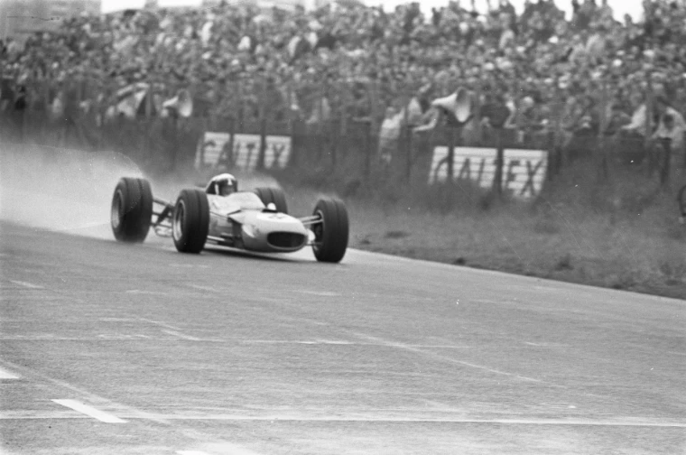 In black and white, a single-seater throws up spray on a wet track beside a crowded bank of spectators (1968).