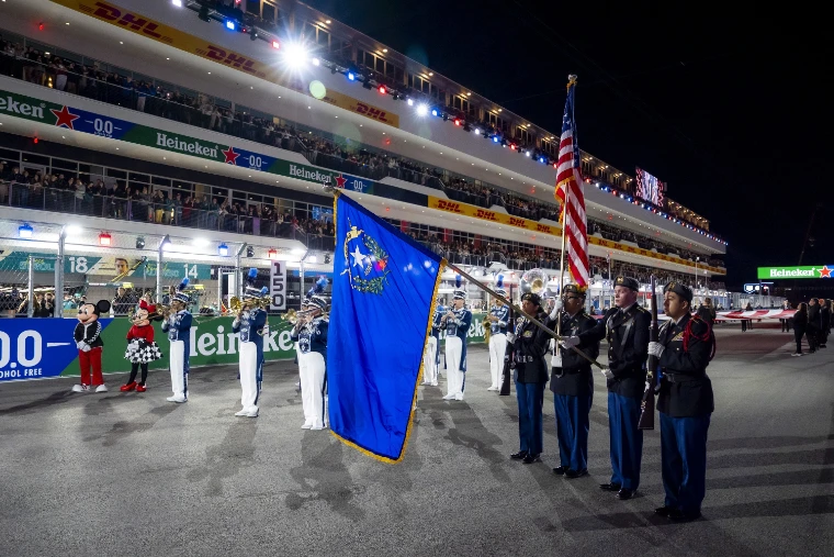 A colour guard holding the American and Nevada flags and a marching band stand on the grid at night before the race, under the floodlit grandstands (2025).