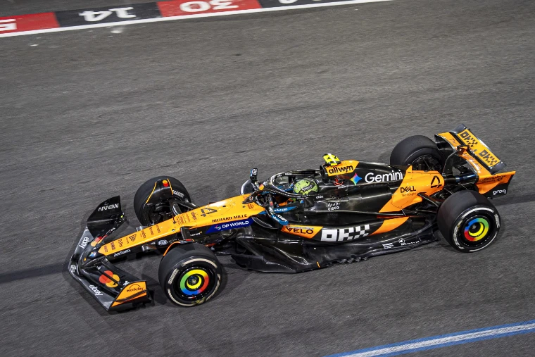 An orange McLaren Formula One car at speed on the floodlit track at night, seen from above with grid numbers painted beside it (2025).