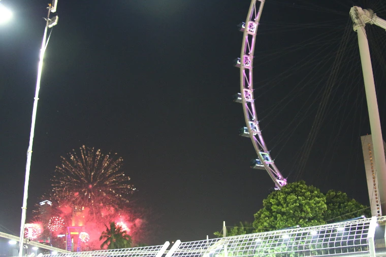 Fireworks bursting red and gold over the city at night beside the lit wheel of the Singapore Flyer, seen past a floodlight mast and the trackside fence (2013).