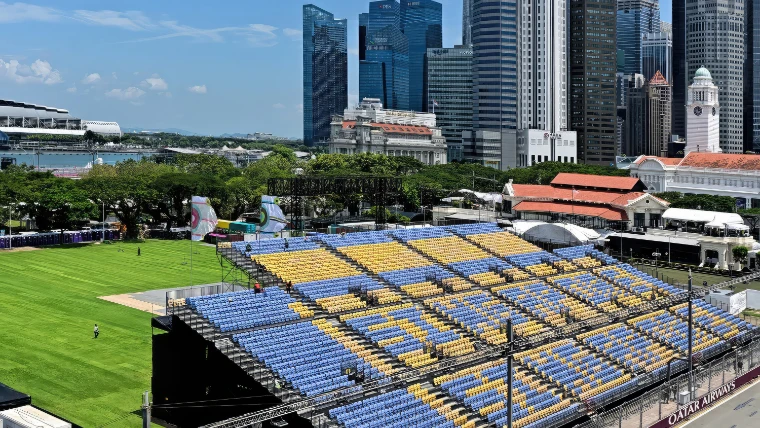Blue and yellow grandstand seating beside the green of the Padang, with colonial buildings, a clock tower and the towers of the business district beyond, in daylight (2023).