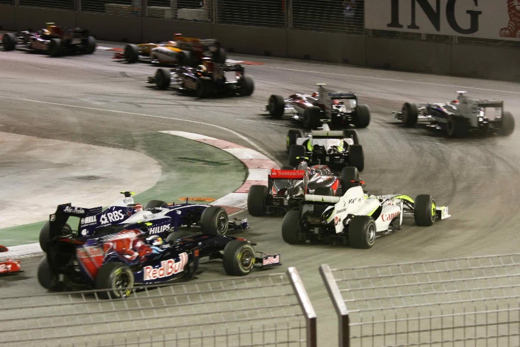 A pack of Formula One cars bunched through the first corner at night under floodlights, seen over a trackside fence (2009).