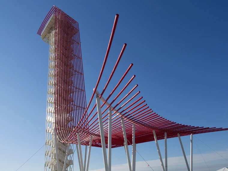 The white observation tower with its sweeping canopy of red steel tubes against a clear blue sky (2012).