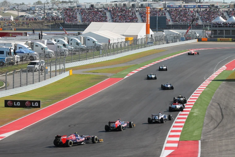 A train of Formula One cars on the first lap through Turns 12 and 13, with packed grandstands and hospitality behind (2012).