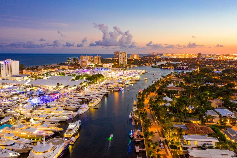 An aerial view at twilight of the illuminated show marinas along the Intracoastal Waterway, the beach and an orange sky beyond (2019).