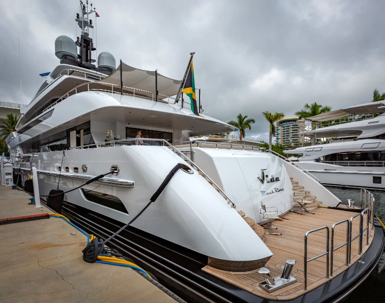 The curving white stern and teak aft deck of a superyacht moored at the dock under a grey sky, palms behind (2019).