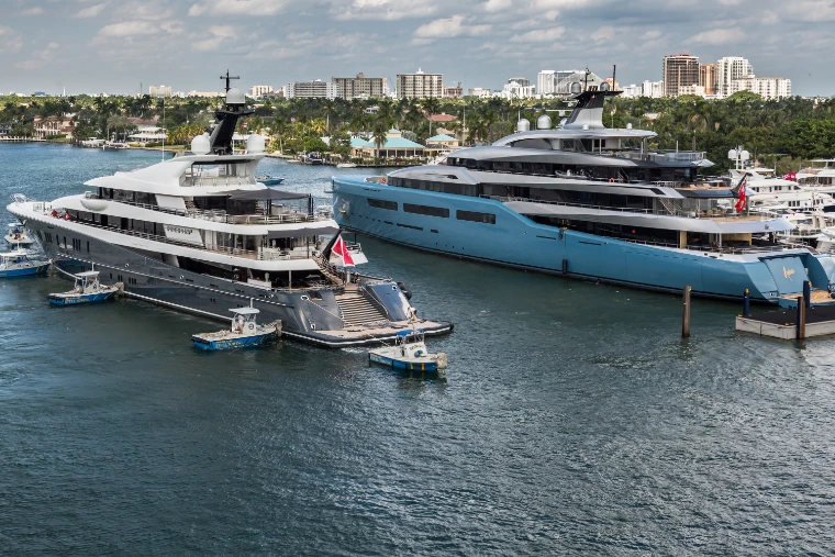Two large superyachts, one grey and one pale blue, berthed side by side at a marina with tenders alongside and the city skyline beyond (2018).