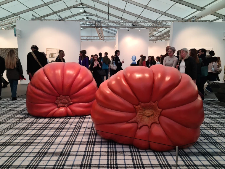 Visitors crowd an aisle of the Frieze London tent around two giant red tomato sculptures on a tartan floor, white booths behind (2022).