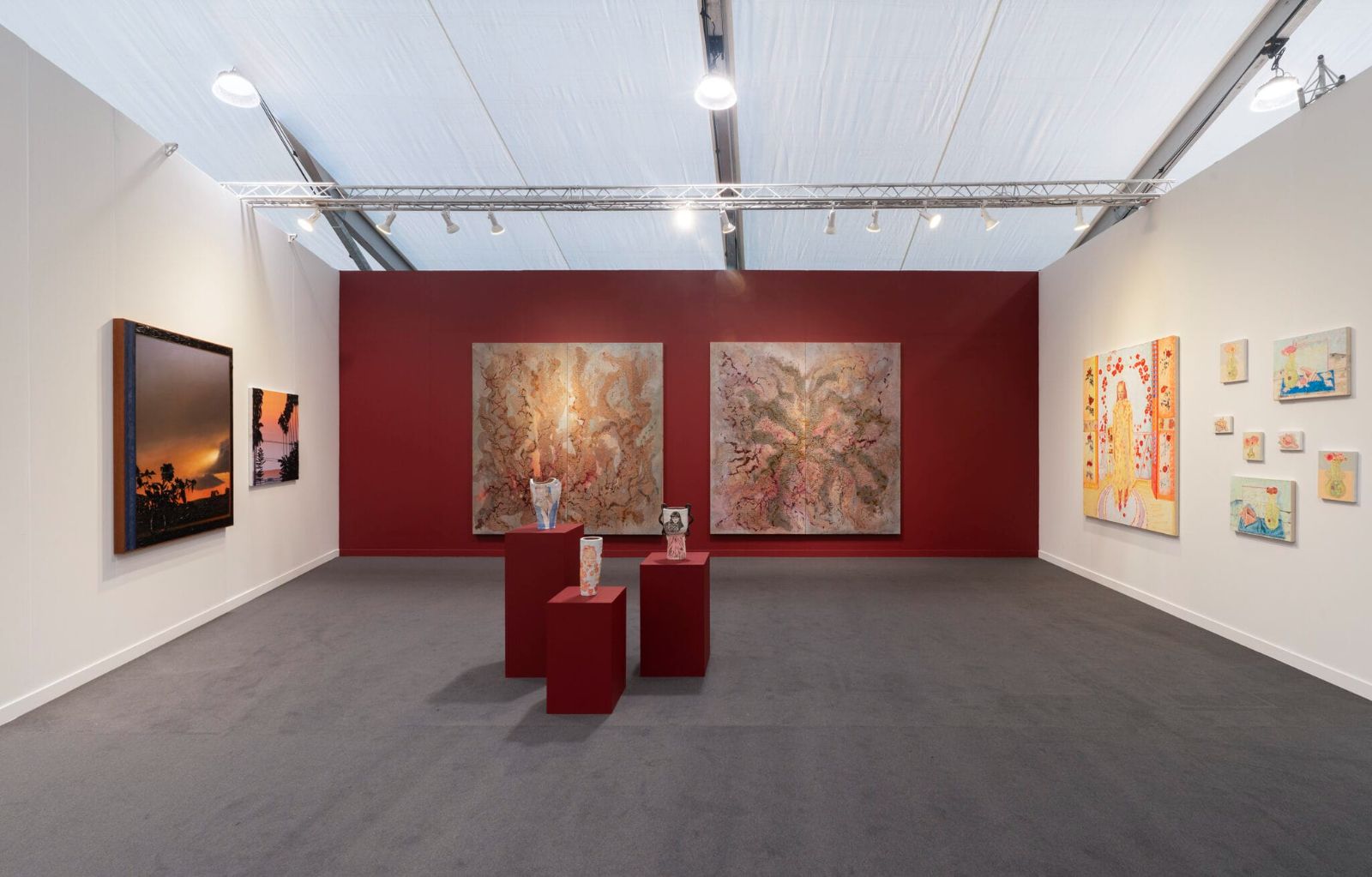 Large paintings on a deep red wall with ceramics on plinths in a gallery booth at Frieze Los Angeles