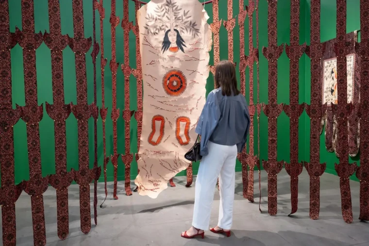 A visitor studies a painted hide hung among patterned textile strips against a green booth wall, in 2025. Frieze New York at The Shed, Hudson Yards, in the season calendar of The Mansa Court