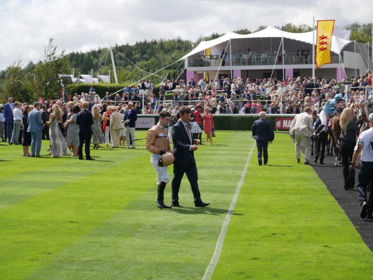 A jockey in silks walks across the striped lawn of the parade ring beside a man in a suit, with crowds and a white canopied stand behind (2021).