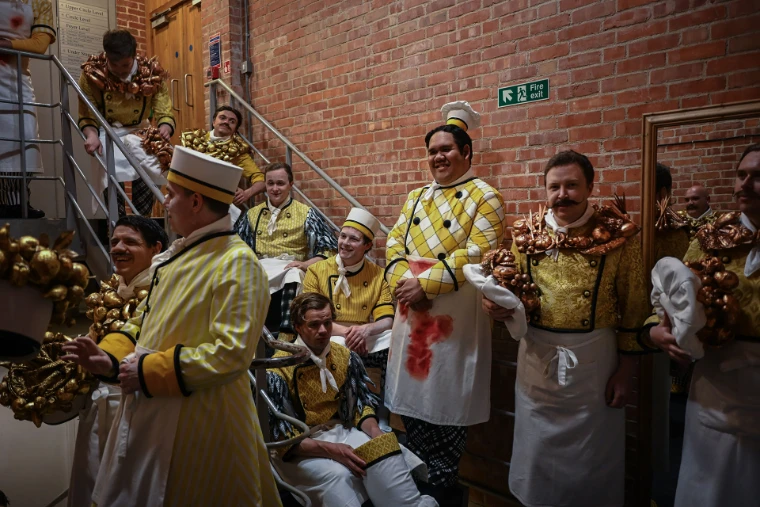 The men’s chorus in yellow cooks’ costume wait on the backstage stair. Glyndebourne Festival Opera, near Lewes in East Sussex, in the season calendar of The Mansa Court