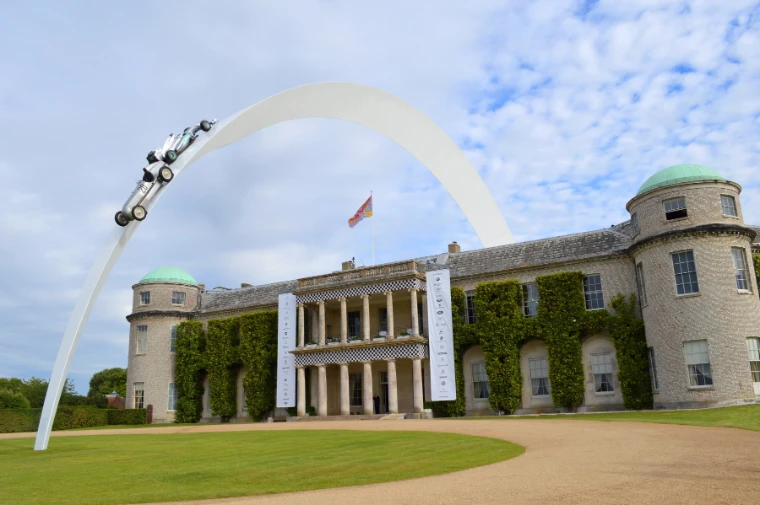 A tall white arched sculpture carrying a racing car curves over the lawn in front of Goodwood House under a blue sky with white clouds (2014).