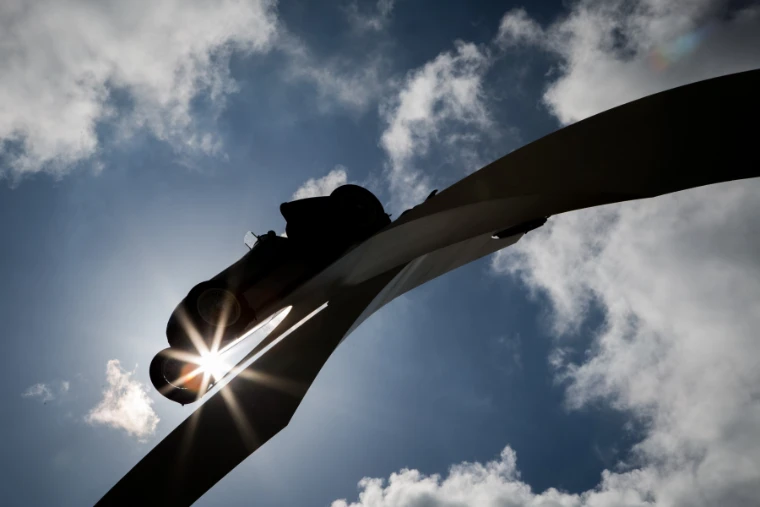 A racing car mounted on the curving arm of the central sculpture is silhouetted against a bright sun and clouds (2014).