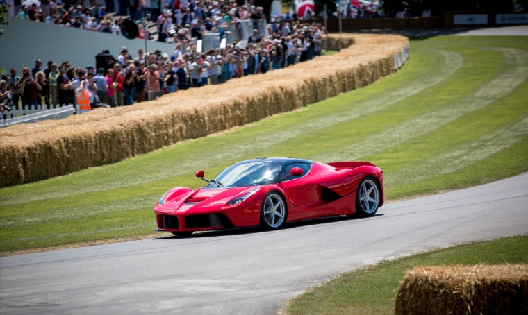 A red Ferrari supercar rounds a bend on the hillclimb course in front of straw bales and a bank of spectators behind a barrier (2014).