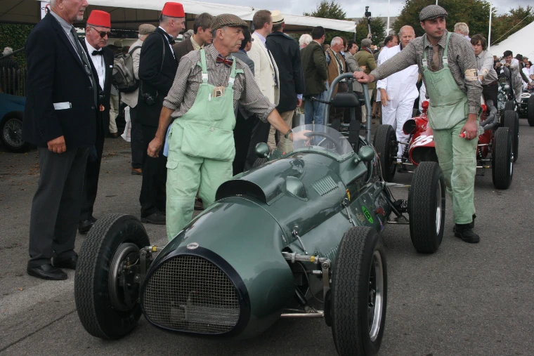 Two mechanics in green period overalls and flat caps push a dark green single-seater racing car past men in jackets, bow ties and fezzes (2011).