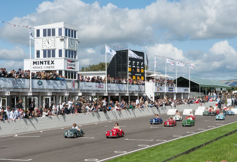 Children race small pedal cars down the start straight past the pits, the clock tower and a crowd along the wall under a summer sky (2017).