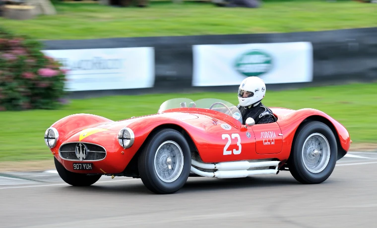 A red 1950s Maserati sports-racing car numbered 23 accelerates away from the chicane, the background blurred with speed (2014).