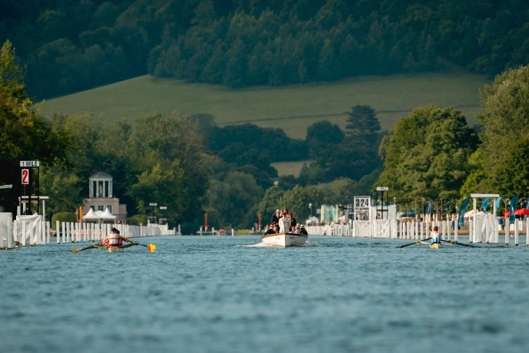 Two crews race up the course with an umpire’s launch behind them, the booms lining the river and the wooded hills beyond.