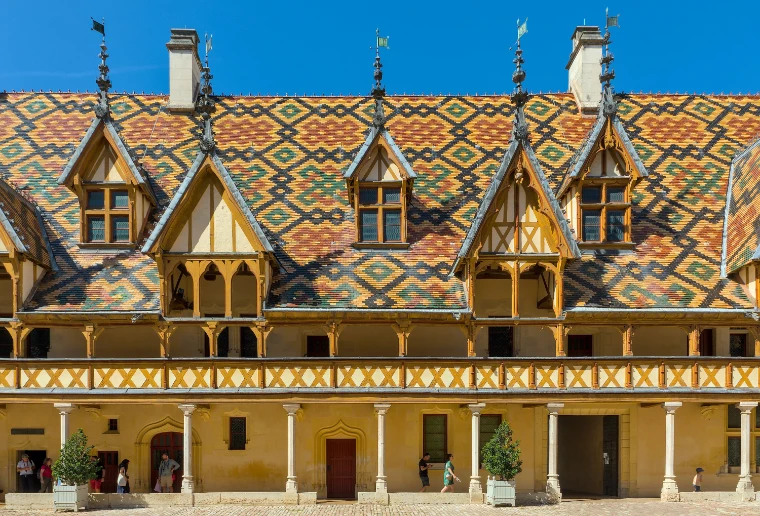 The courtyard of the Hôtel-Dieu, its steep roofs of red, brown, yellow and green glazed tiles and gabled dormers rising above a timbered gallery, under a blue sky (2025).