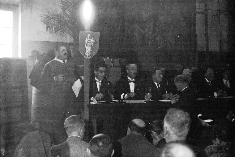 Black-and-white photograph of the Hospices de Beaune wine sale, a crier standing beside a shield of the Virgin and Child while men in evening dress sit at the sale table with a candle burning, the bidders seated below (1932).