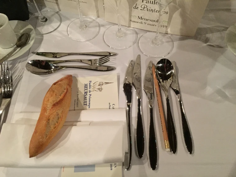 A single place at a Paulée table, with a row of wine glasses, silver cutlery, a roll of bread and a printed menu on a white cloth (2019).
