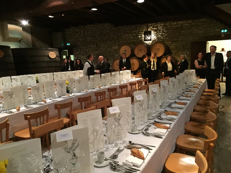 Guests in jackets gather by the barrels at the end of the cuverie of the Château de Meursault beside long tables laid with glasses and menus (2019).