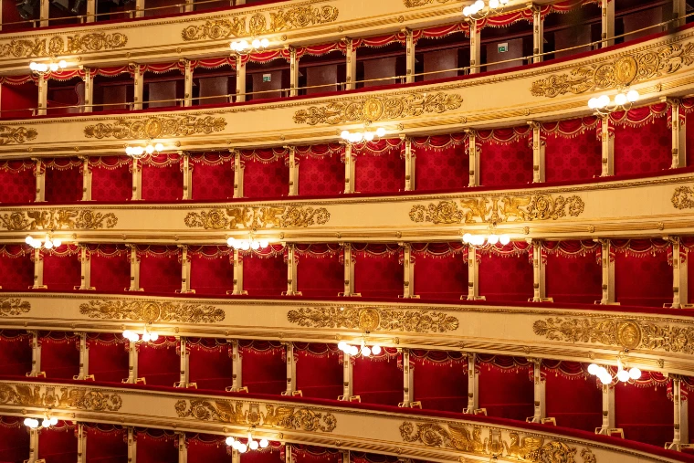 Tiers of red-velvet boxes with gilded balustrades and small lamps filling the frame inside La Scala (2023).