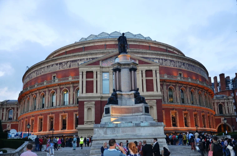 The red-brick Royal Albert Hall at dusk with its lights coming on, a statue on a tall stone pedestal before it and concertgoers gathering on the steps below (2014).