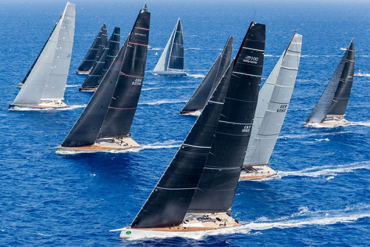 A fleet of maxi yachts under dark and silver sails racing across open blue water, the class that contests Les Voiles de Saint-Barth, a mid-April engagement in the season calendar of The Mansa Court