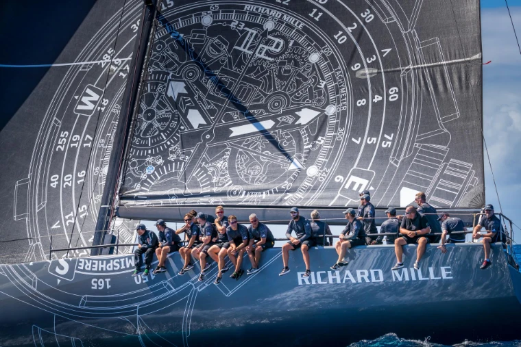 The crew of a maxi yacht lined along the rail beneath a mainsail printed with a watch dial at Les Voiles de Saint-Barth, a mid-April engagement in the season calendar of The Mansa Court