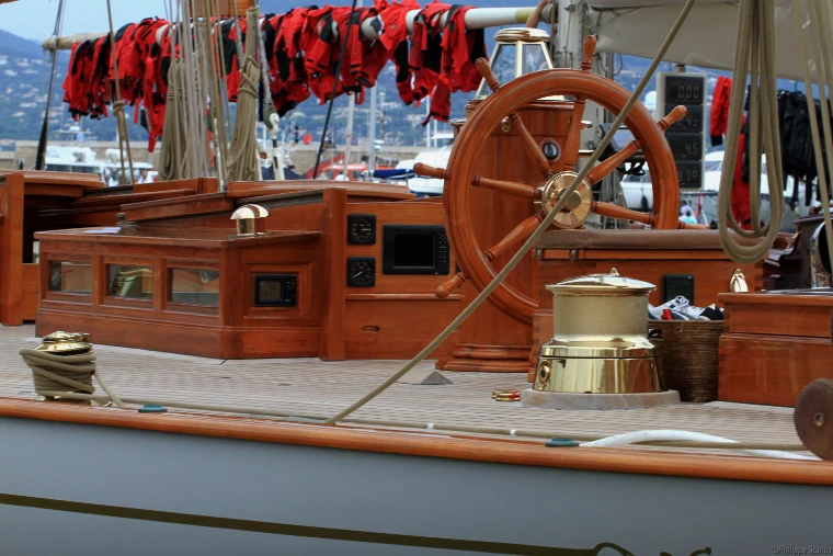 The varnished wooden wheel, brass winch and teak deck of a classic yacht moored in Saint-Tropez, with red life jackets hung along the rigging (2014).
