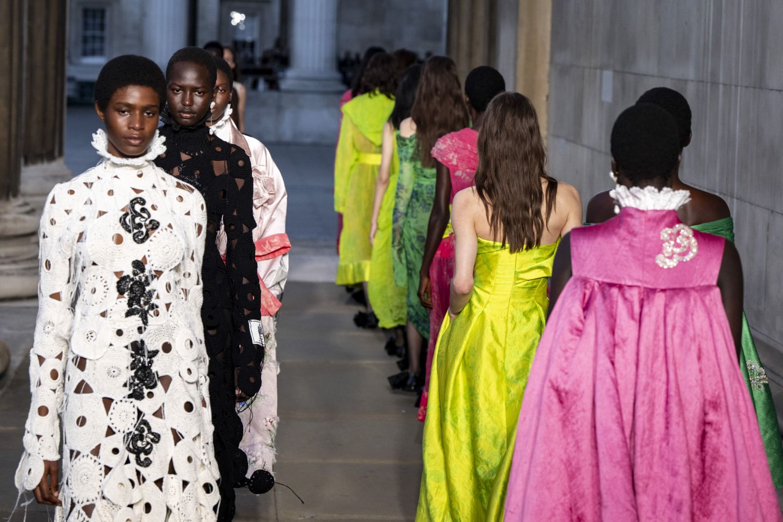 Models in acid green, magenta and cut lace walking a runway through a stone colonnade at London Fashion Week