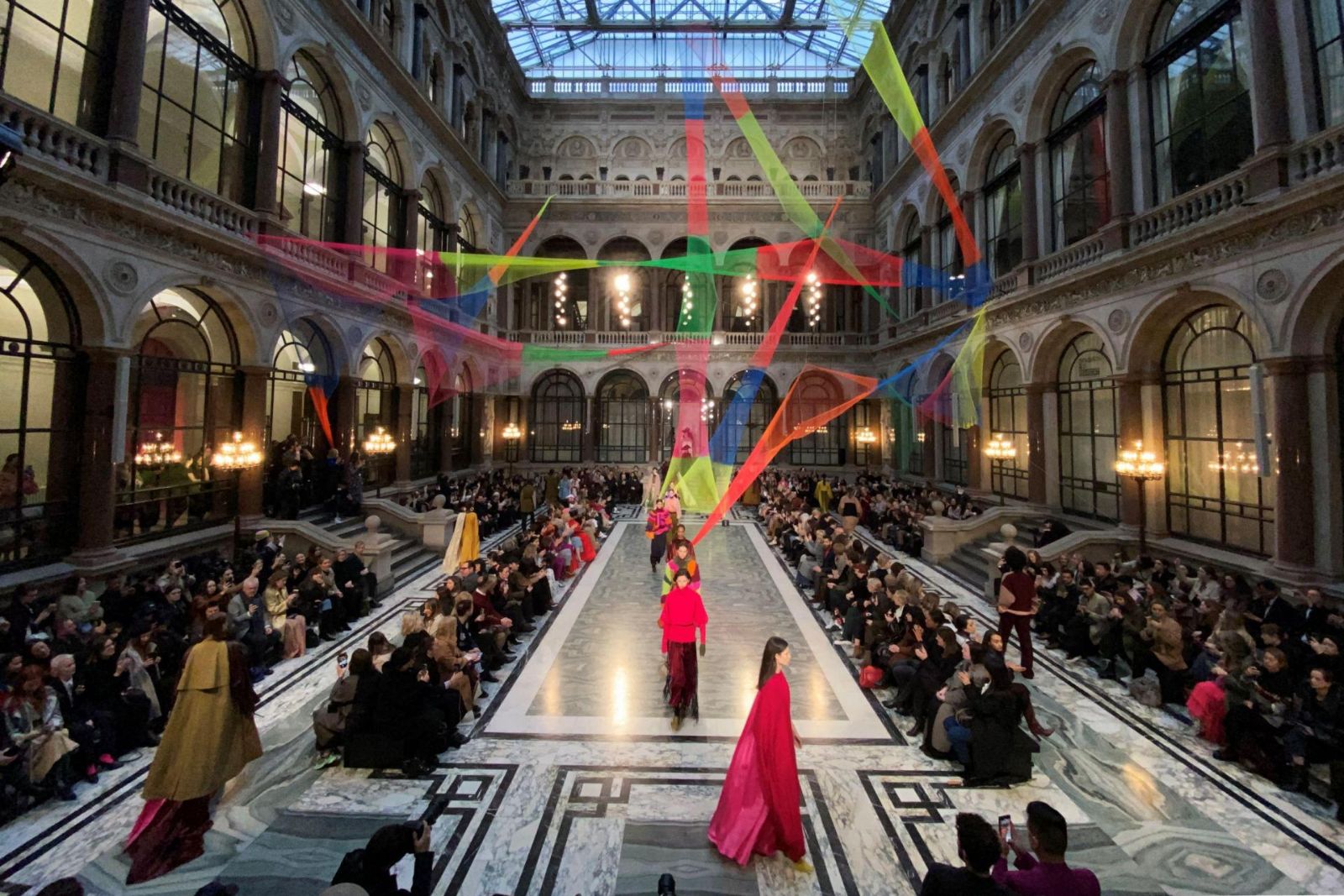 Models walking a marble runway beneath a coloured ribbon installation in a glazed Victorian atrium at London Fashion Week