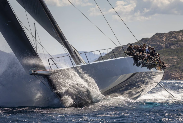 A grey-hulled maxi yacht heels hard through spray with its crew lined along the windward rail and a rocky shore behind (2024).