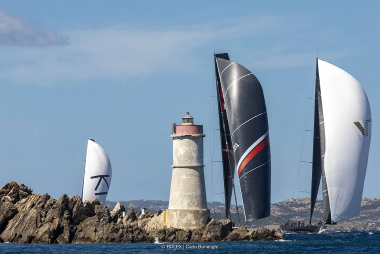 Three maxi yachts under spinnaker sail past a small stone lighthouse on a granite reef (2024).
