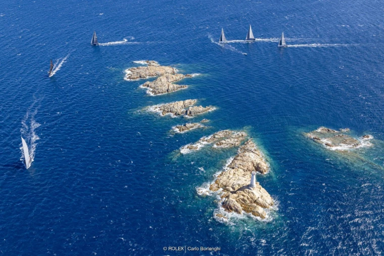 An aerial view of maxi yachts racing past a chain of granite islets, one with a white lighthouse, in deep blue water (2024).
