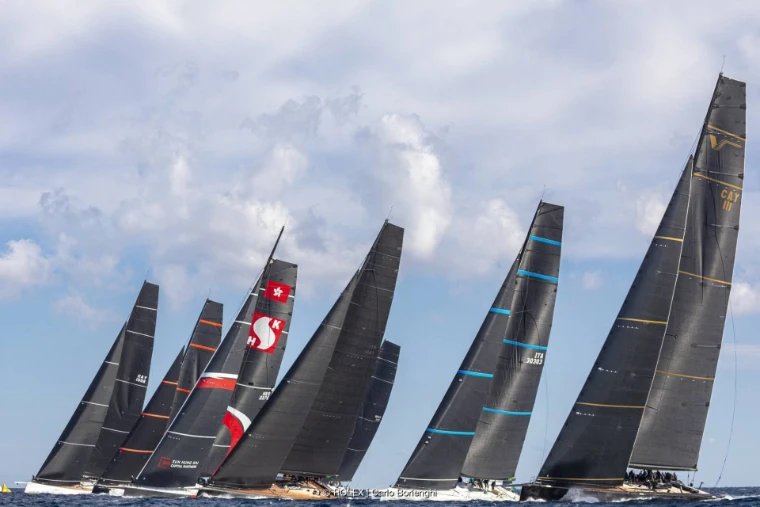 A tightly bunched line of maxi yachts with dark racing sails heads upwind under a cloudy sky (2024).