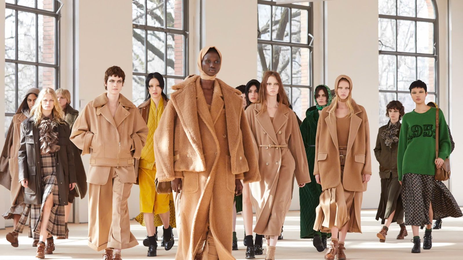 Models in camel coats, shearling and hooded knits walking through a daylit hall of tall arched windows at Milan Fashion Week
