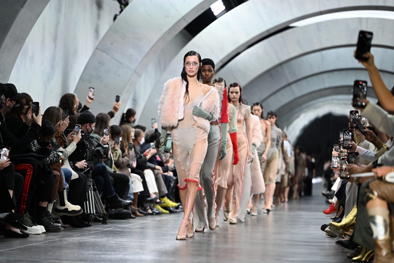 Models in blush chiffon and shearling walking a runway through a vaulted concrete hall as the front row films at Milan Fashion Week