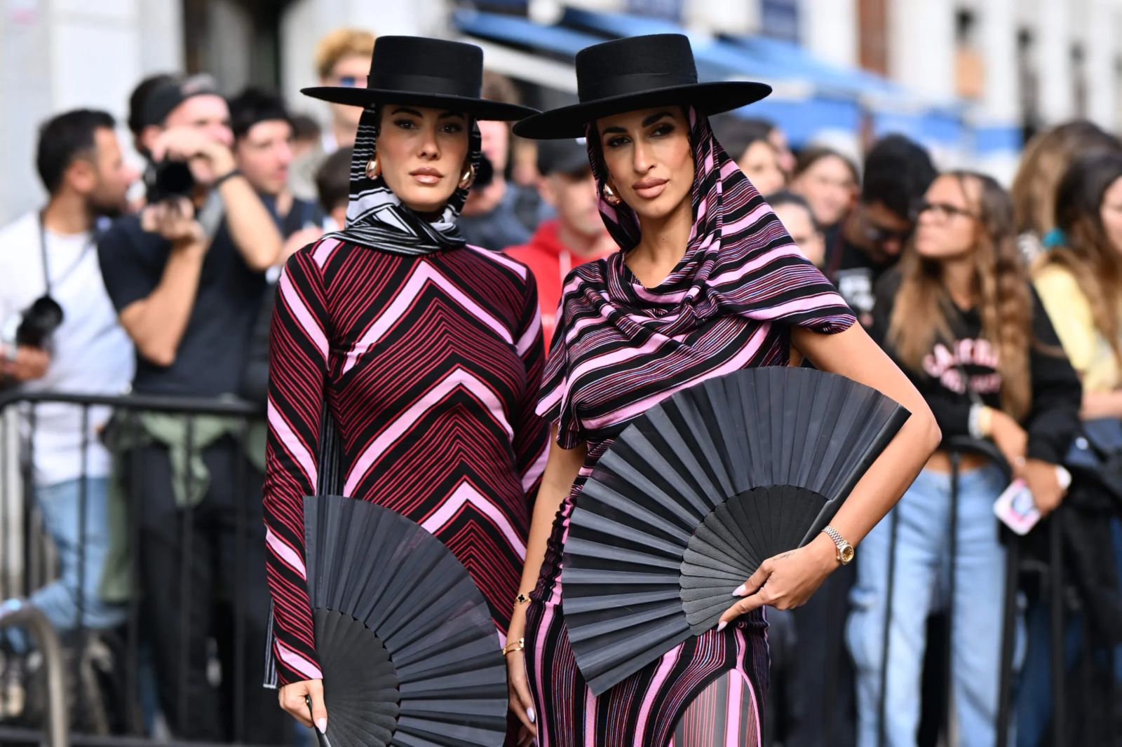 Two guests in pleated stripes, wide-brimmed hats and hand fans photographed outside a show at Milan Fashion Week
