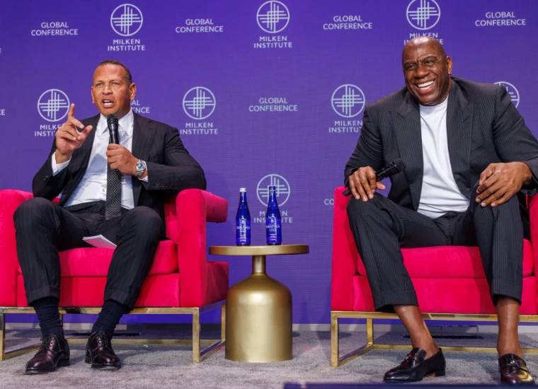 Two speakers in pinstripe suits share the stage in red velvet chairs before the violet Global Conference backdrop. The Milken Institute Global Conference at The Beverly Hilton, Beverly Hills, in the season calendar of The Mansa Court