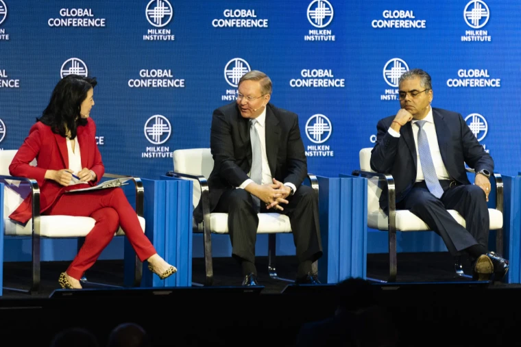 A moderator in red questions two panellists on the main stage beneath the Global Conference screen. The Milken Institute Global Conference at The Beverly Hilton, Beverly Hills, in the season calendar of The Mansa Court