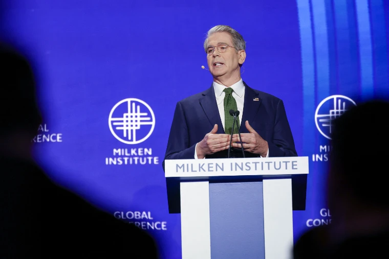 A speaker addresses the hall from the Milken Institute lectern before the blue conference screen, in 2025. The Milken Institute Global Conference at The Beverly Hilton, Beverly Hills, in the season calendar of The Mansa Court
