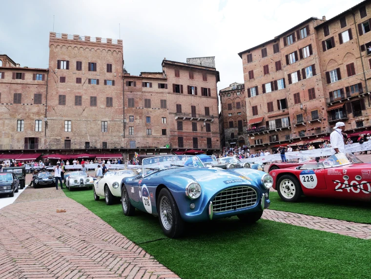A blue British roadster waits on the green carpet in the Campo, the tall brick palaces of Siena behind. The Mille Miglia, from Brescia to Rome and back, in the season calendar of The Mansa Court