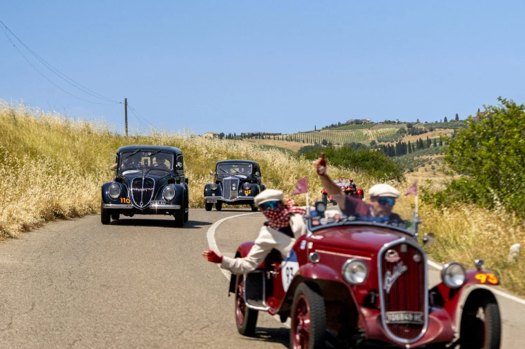 A red open two-seater and two black saloons wind along a country road through the Tuscan hills, the crew waving, in 2025. The Mille Miglia, from Brescia to Rome and back, in the season calendar of The Mansa Court