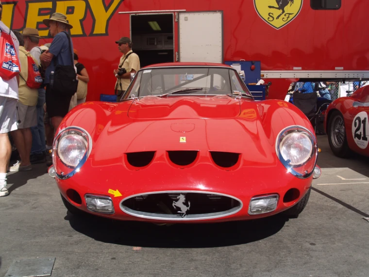 The front of a red Ferrari 250 GTO in the paddock, with team members and visitors standing beside a red Ferrari transporter (2007).