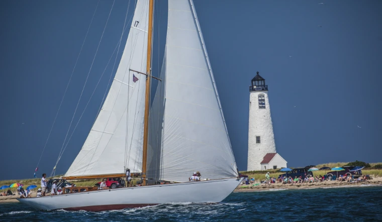 A classic white sloop under full sail heels past a white stone lighthouse while people on the beach watch from under colourful umbrellas (2015).