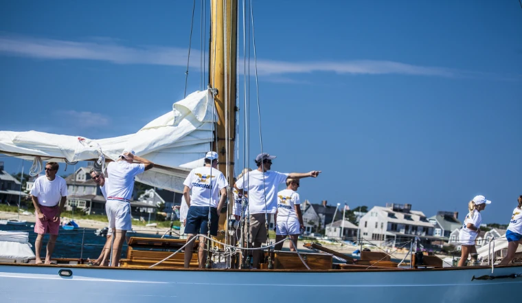 Crew members in white shirts and shorts handle sails on the varnished deck of a classic yacht, one pointing ahead, with shingled waterfront houses behind (2015).