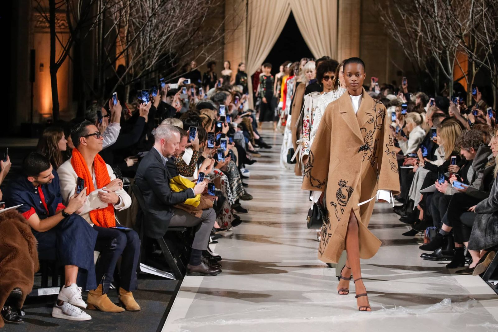 An embroidered camel coat on the runway as the front row raises its phones at New York Fashion Week